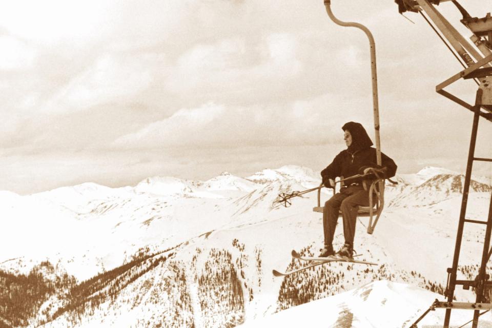 Vintage photograph of a skier riding an early chairlift at Arapahoe Basin, suspended above a snowy mountain landscape during the early days of lift-served skiing.